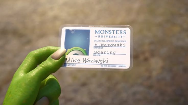 Close-up of a Monsters University student ID card for Mike Wazowski, held in a green monster hand, showing his photo, name and “Scaring” as his major.