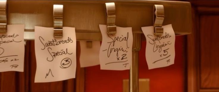 Close-up of handwritten restaurant order tickets clipped above the pass in a kitchen, with notes like “Sweetbreads Special” and “Special Linguini” hanging in a row